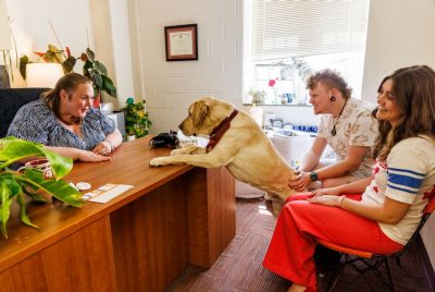 A yellow Labrador retriever wearing a maroon bandana puts its front paws on a desk to interact with a smiling woman seated behind it, while two students sitting nearby pet the dog and laugh. The setting is a warmly lit office with plants, a diploma on the wall, and sunlight streaming through a window. 
