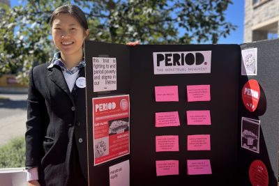  A smiling student stands outdoors beside a table with a trifold poster showing questions about menstrual periods.