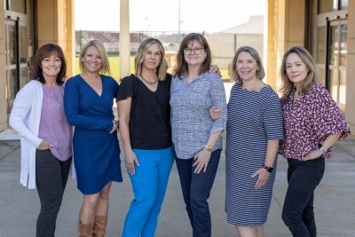 Six women stand smiling alongside each other in the breezeway entrance of a building, with an athletic field in the background.